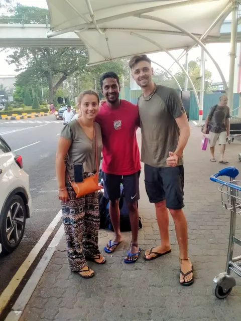 Two guests with their driver-guide on the pavement outside the airport, a luggage trolley behind them