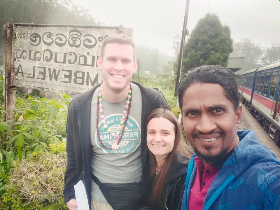 Two guests and their driver-guide taking a selfie by the Ambewela station sign, with a train waiting behind