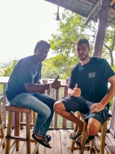 A guest and their driver-guide giving a thumbs-up from bar stools on a wooden veranda