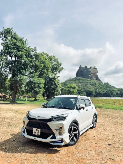 A white car parked on a dirt clearing, with Sigiriya Rock rising above the trees behind it