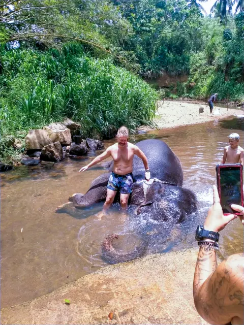 A guest sitting on an elephant’s back in a shallow river while someone takes a photo