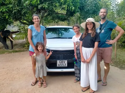 Two adults and three children standing in front of a white car on a sandy clearing