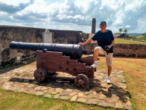 A guest resting a foot on the carriage of an old cannon on a fort rampart, with a clock tower in the distance