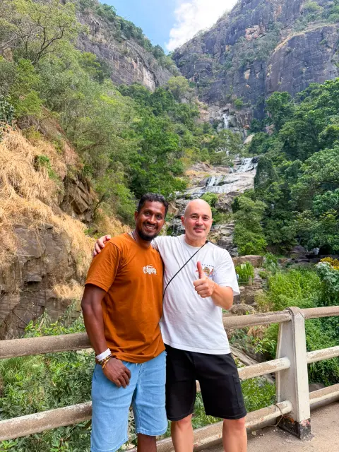 A guest and their driver-guide at a railing above a forested gorge, with a waterfall in the distance