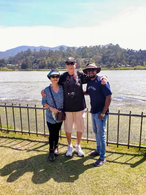 Two guests and their driver-guide at a lakeside railing with hills beyond