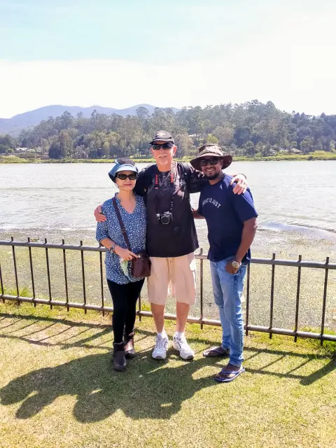 Two guests and their driver-guide on a lawn beside a lake, with wooded hills across the water