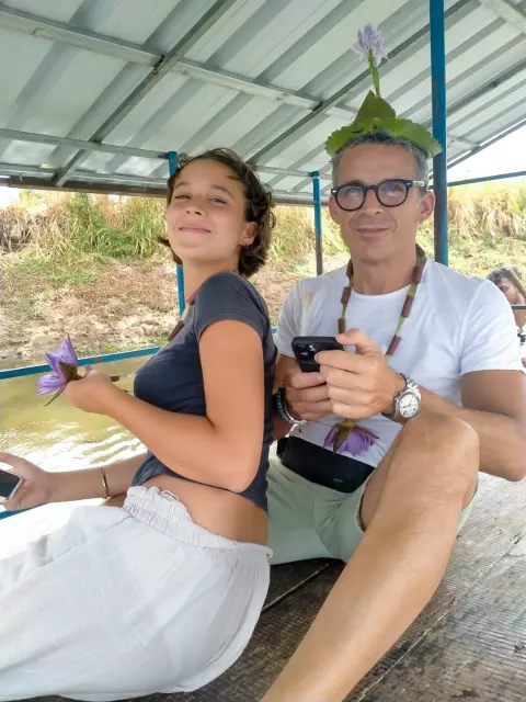 Two guests on a canopy boat, one holding a lotus flower and the other wearing a lotus-leaf hat