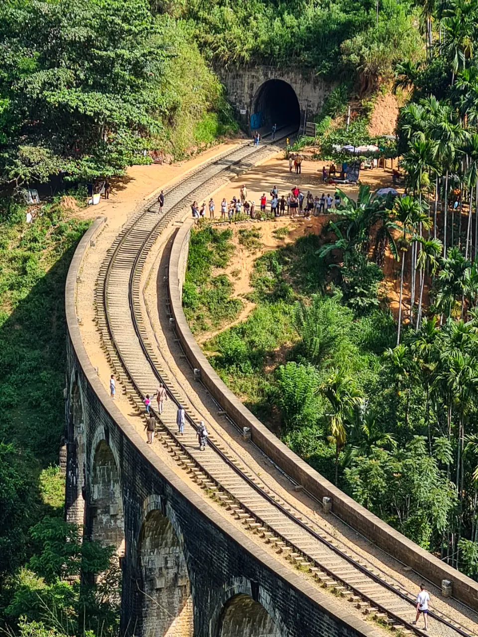 Looking down on the curving Nine Arches Bridge as the railway runs into a tunnel through the jungle, with people walking along the tracks