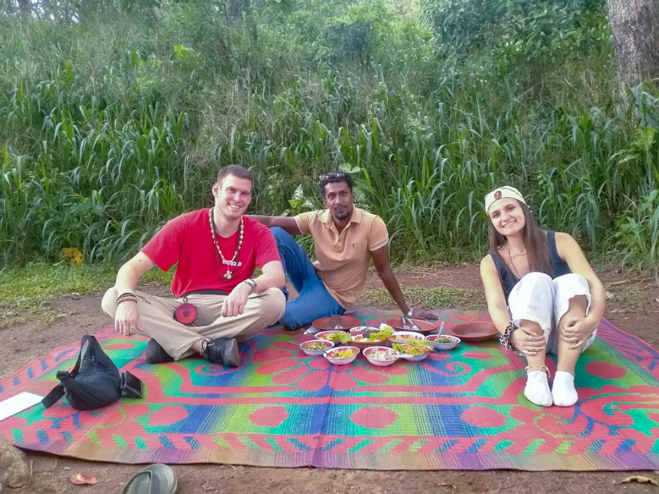 Two guests and their driver-guide sitting on a colourful mat with a spread of dishes, tall crops behind