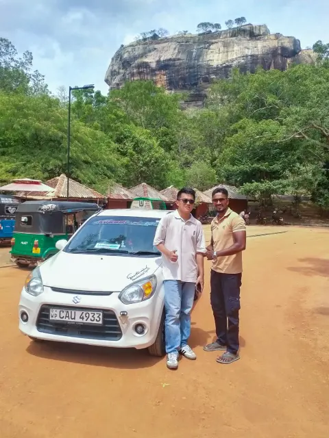 Sigiriya Rock rising above the trees behind two men standing beside a small white car, with tuk-tuks parked nearby