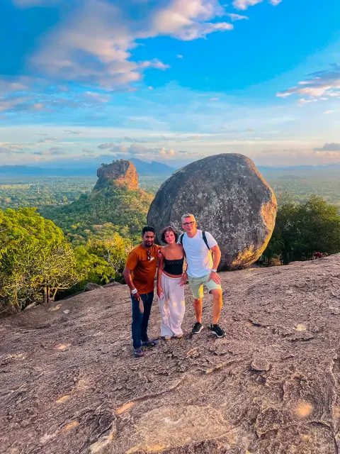 Three people standing on a bare rock summit beside a huge boulder, with Sigiriya Rock rising from the forest behind them