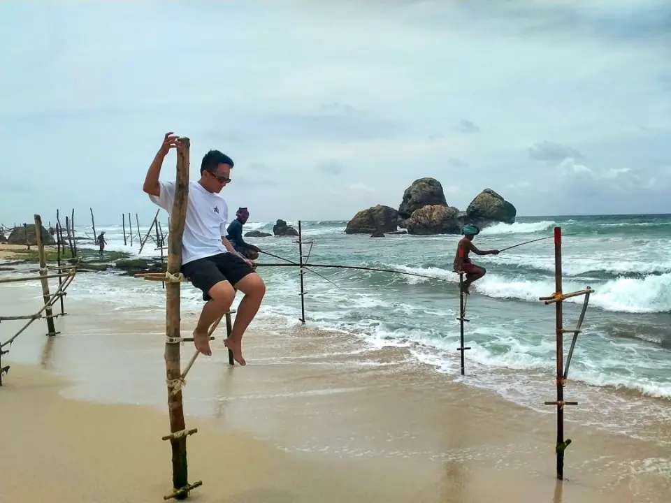 A guest perched on a stilt-fishing pole at the water’s edge, with waves breaking around rocks behind