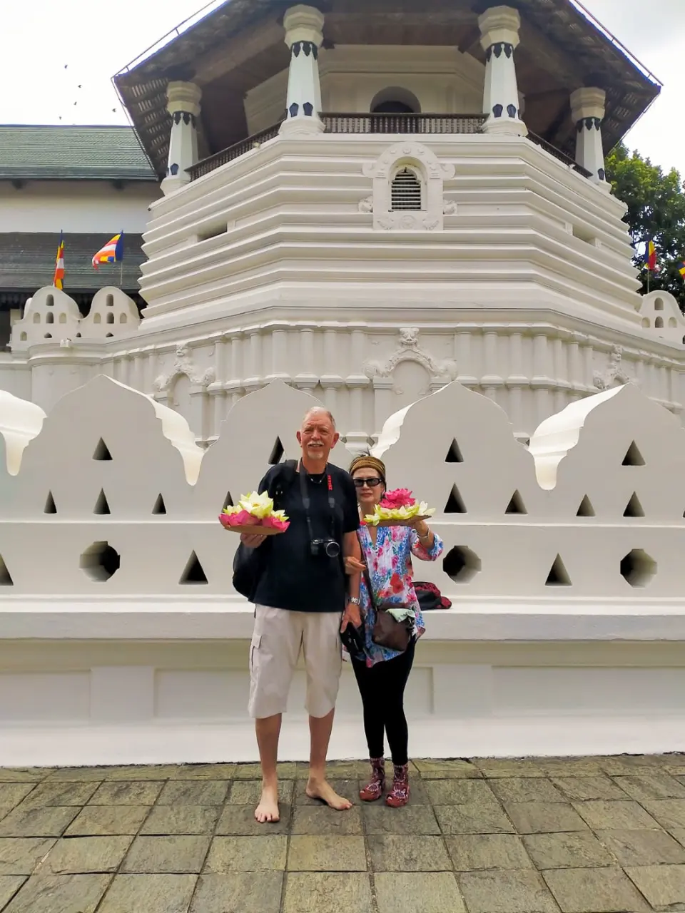 Two guests holding lotus-flower offerings in front of the white octagonal pavilion of the Temple of the Sacred Tooth Relic in Kandy