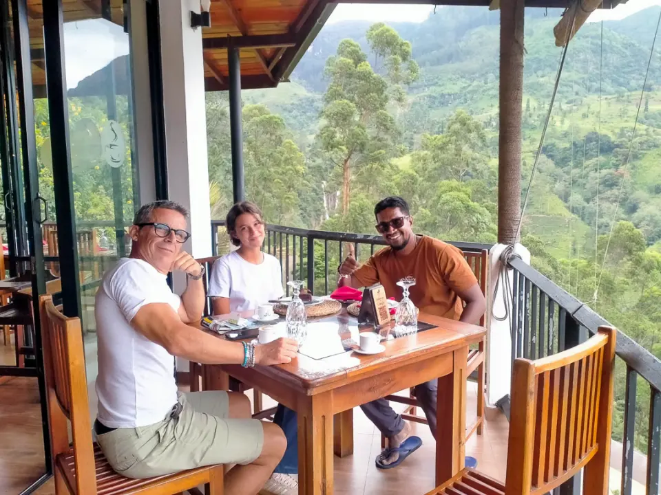 Two guests and their driver-guide at a table on a wooden terrace looking out over green hills
