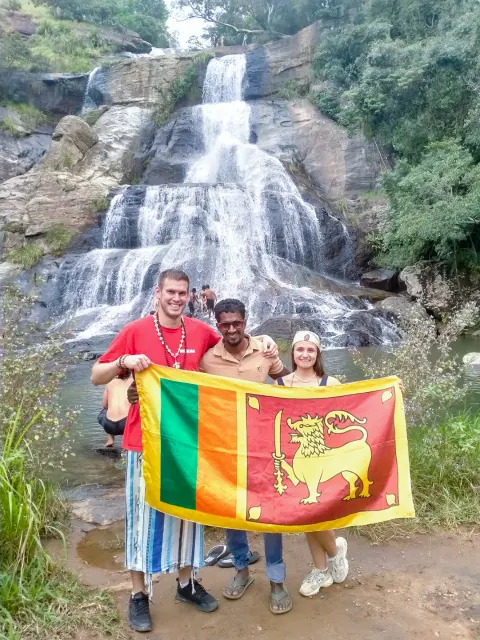 Guests and their driver-guide holding up a Sri Lankan flag in front of a tall waterfall