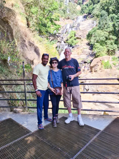 Two guests and their driver-guide on a viewing platform below a tall waterfall