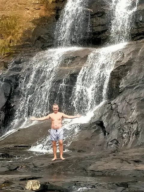 A guest standing with arms outstretched on the rocks beneath a cascading waterfall