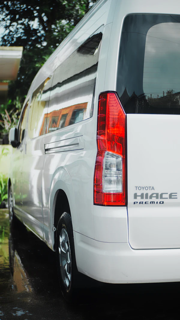 The rear of a clean white Toyota HiAce van parked beside greenery